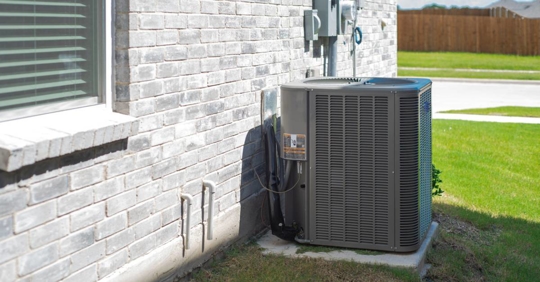 central air conditioning condenser unit mounted on concrete slab beside brick house wall